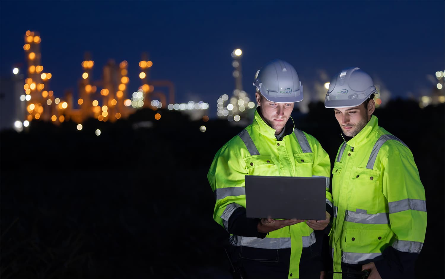 Two site workers in high-vis looking at a laptop at night with a city skyline behind